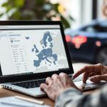 Un homme compare des annonces de voitures d’occasion sur un ordinateur portable, dans un bureau baigné de lumière naturelle. Une voiture est visible à l’arrière-plan et une carte d’Europe apparaît de façon floue.