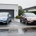 Deux voitures compactes comparables, l’une électrique branchée à une borne domestique et l’autre essence dans une station-service, photographiées de manière réaliste.