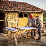 Dans quel ordre faut-il réaliser les travaux de rénovation d’une maison ? Maison ancienne en rénovation avec toiture réparée, murs ouverts et deux artisans examinant les plans du chantier.
